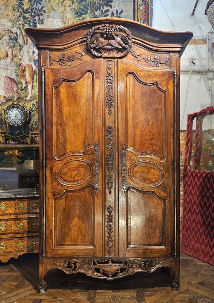 An 18th-century walnut wedding armoire. Beautiful, animated cornice featuring a pediment carved with a laurel wreath framed by two doves on a basket, with ribbons and olive branches.On the door frame, there are additional symbols of a successful union: the arrow of love, an urn and flowers for fertility, and oak leaves for strength. The lower rail is openwork, carved with an urn and foliage. The piece stands on cabriole feet with scrolls.