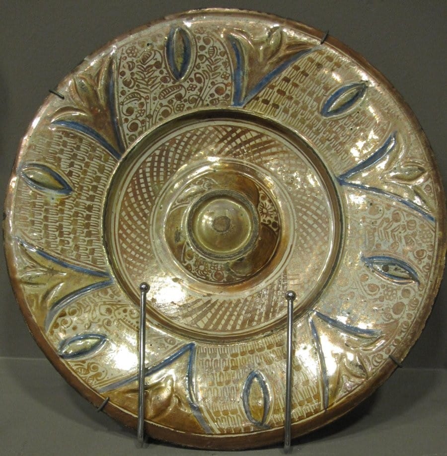 This 16th-century Hispano-Moresque lustreware dish features a raised central boss. The broad rim is decorated with foliage highlighted in blue against a background strewn with alternating solfas and tiny flowers.
